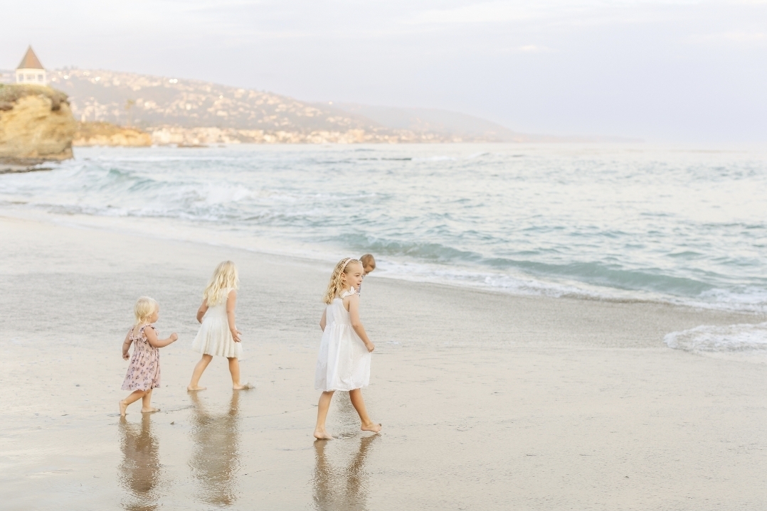 Three young children walking barefoot along the shoreline of Laguna Beach at sunset, captured in soft, golden light.
