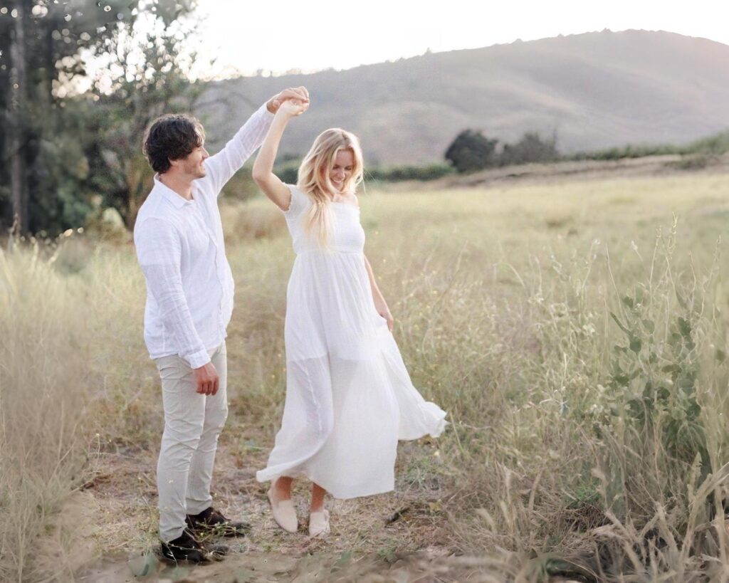 Playful couple dancing together in an open flower field at golden hour, with hills in the background — photographed by Terialina Photography, a couples photographer in Orange County.